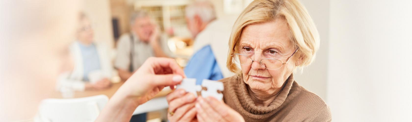 Starsza kobieta w okularach trzyma dwa puzzle i próbuje je dopaswać. Ktoś jej pomaga.