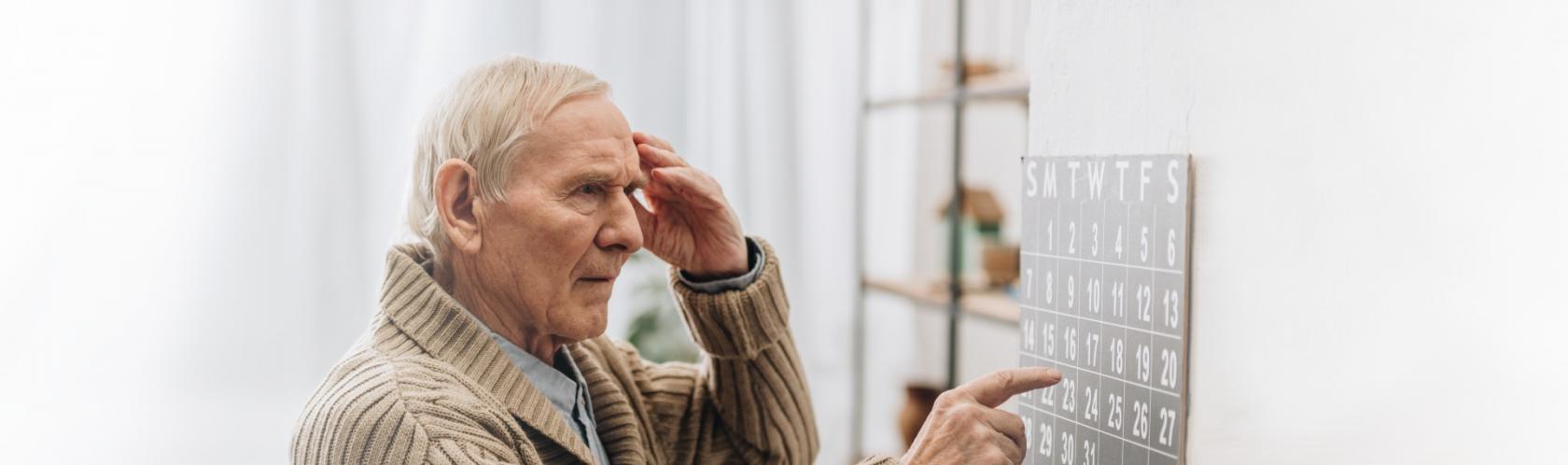 Starszy mężczyzna patrzy na kalendarz i wskazuje datę. Drugą ręką dotyka skroni. Wygląda na zdezorienrowanego.
