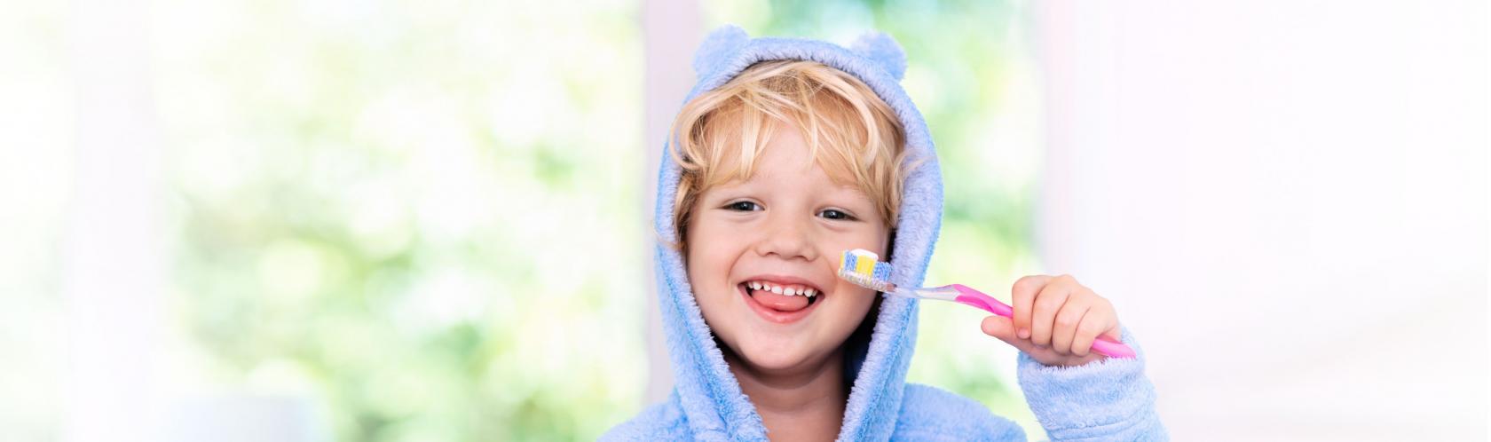 Kilkuletni chłopczyk w szlafroku trzyma szczoteczkę do zębów. Wystawia język, widać mu zęby mleczne.