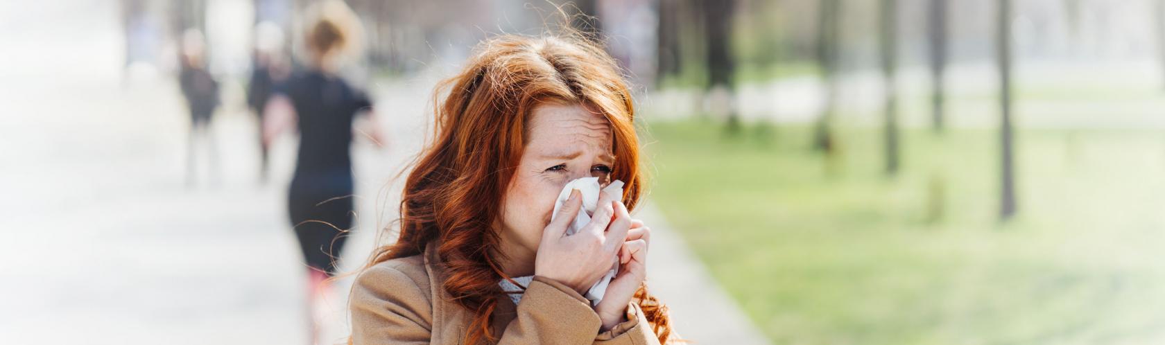 Kobieta, która jest uczulona na pyłki, kicha i zasłania nos chusteczką. Stoi na chodniku, ma rude włosy.