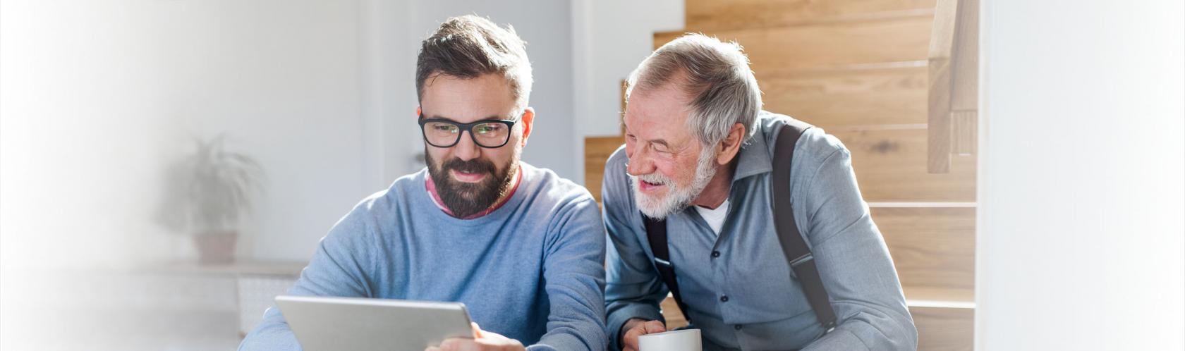 Dorosły syn z ojcem siedzą na schodach w domu. Syn pokazuje coś ojcu na tablecie, senior ma w dłoniach kubek.