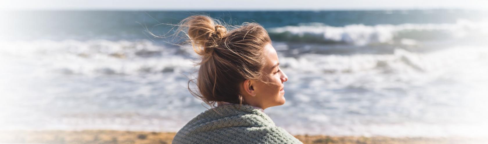 Młoda kobieta w blod włosach spiętych w koczek siedzi na plaży. Patrzy w stronę morza, słońce oświetla jaj twarz. Na ramionach ma koc.