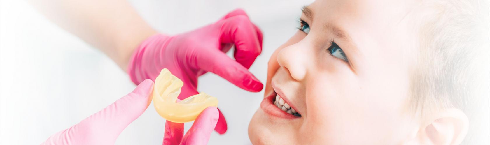 Chłopiec podczas wizyty u ortodonty. Lekarz w różowych rękawiczkach zbliża wycisk do jego ust.