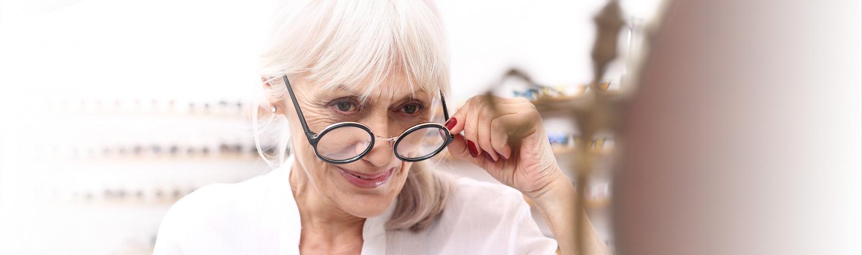 Starsza pani przymierza okulary u optyka. Ma siwe włosy i czerwone paznokcie.