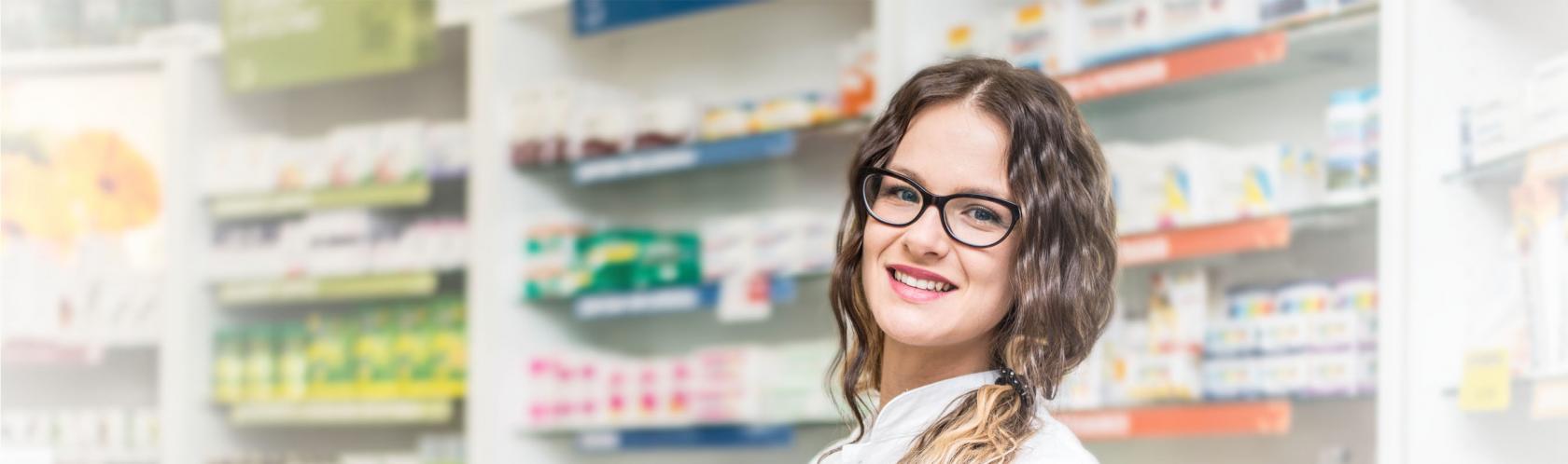 Uśmiechnięta farmaceutka w aptece. Młoda dziewczyna ma długie, falowane włosy upięte w kucyk na boku. Ma okulary.