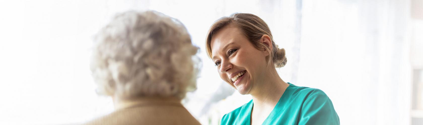 Przyjazna pielęgnierka uśmiecha się do starszej pani i trzyma jej rękę na plecach.