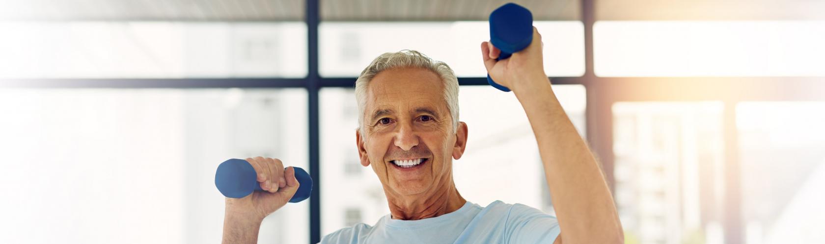 Starszy mężczyzna ćwiczy. Podnosi ciężarki na wysokość głowy. Ma siwe włosy, błękitny t-shirt. Uśmiecha się.