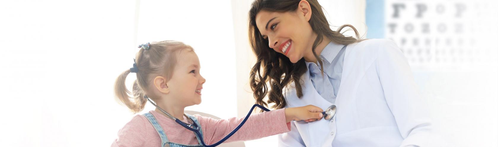Mała dziewczynka podczas wizyty kontrolnej u lekarza. Na kolanach ma misia, na szyi stetoskop, którym bada lekarkę. Obie się uśmiechają