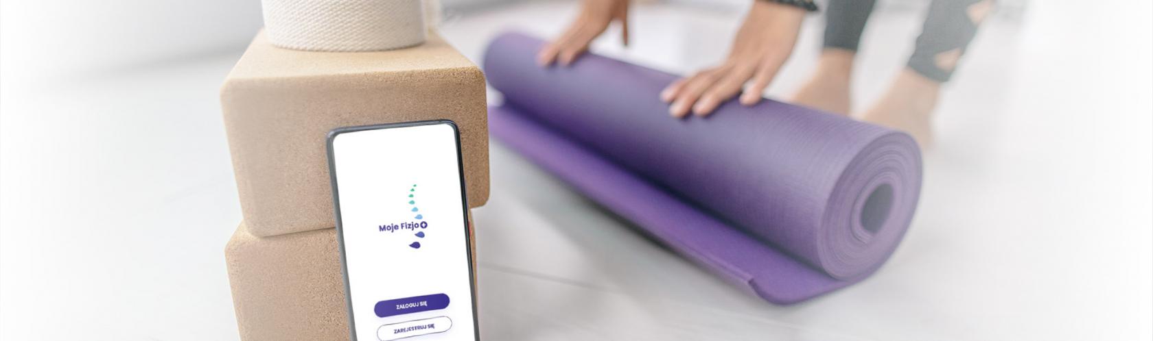 Smartfon z aplikacją Moje Fizjo na ekranie. W tle ręce rolujące matę do ćwiczeń.