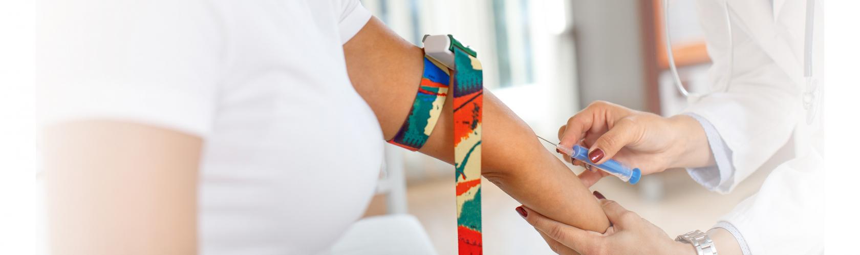 Kobieta podczas oddawania krwi. Na ramieniu ma opaskę uciskową. Lekarka wbija jej igłę. 