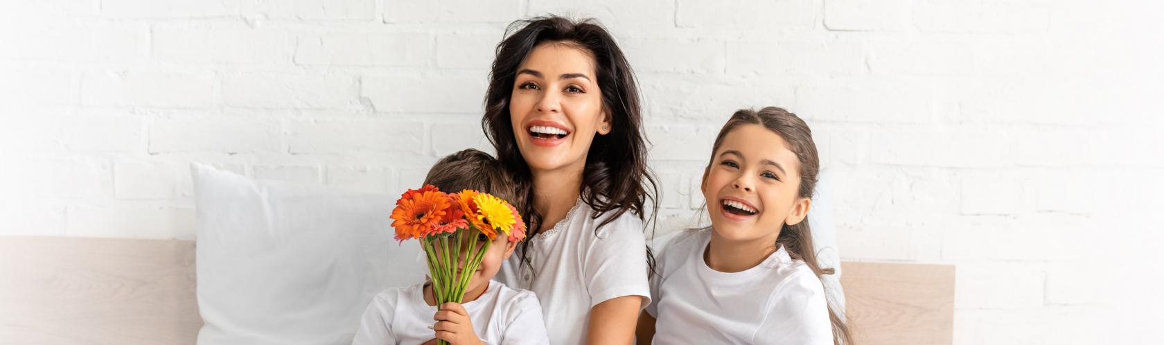 Małoda matka z dwójką kilkuletnich dzieci siedzi na kanapie. Wszyscy się uśmiechają. Chłopiec zakrywa twarz bukietem pomarńczowych kwiatów.