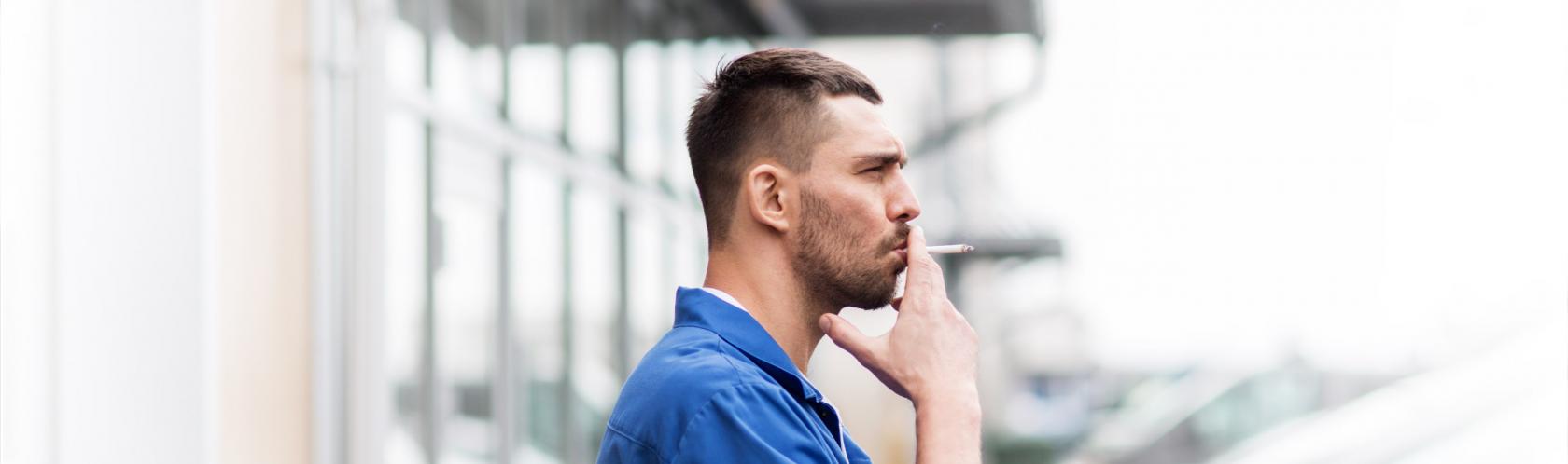 Profil mężczyzny, który stoi przed warsztatem i pali papierosa. Jest ubrany w kombinezon, ma lekki zarost.