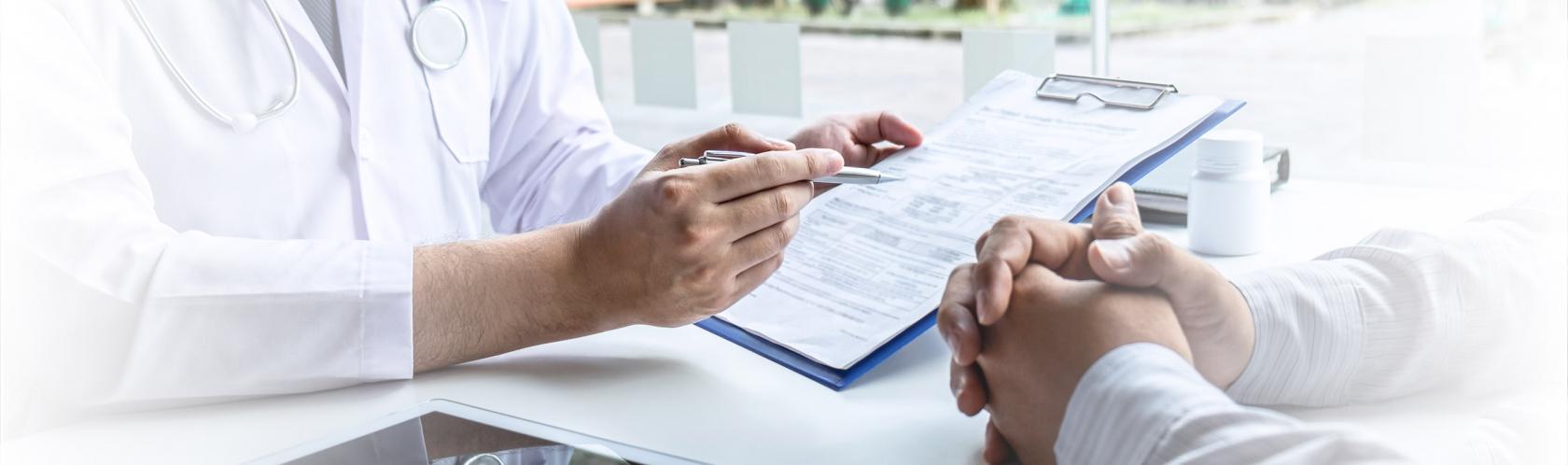 Lekarz omawia wyniki z pacjentem. Pokazuje mu jakiś wydruk. Zbliżenie na ręce na biurku.
