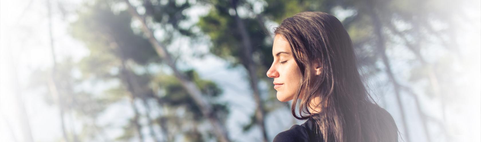 Kobieta stoi nad morzem, w tle rosną sosny. Ma zamknięte oczy, twarz wystawia do słońca. Delektuje się chwilą