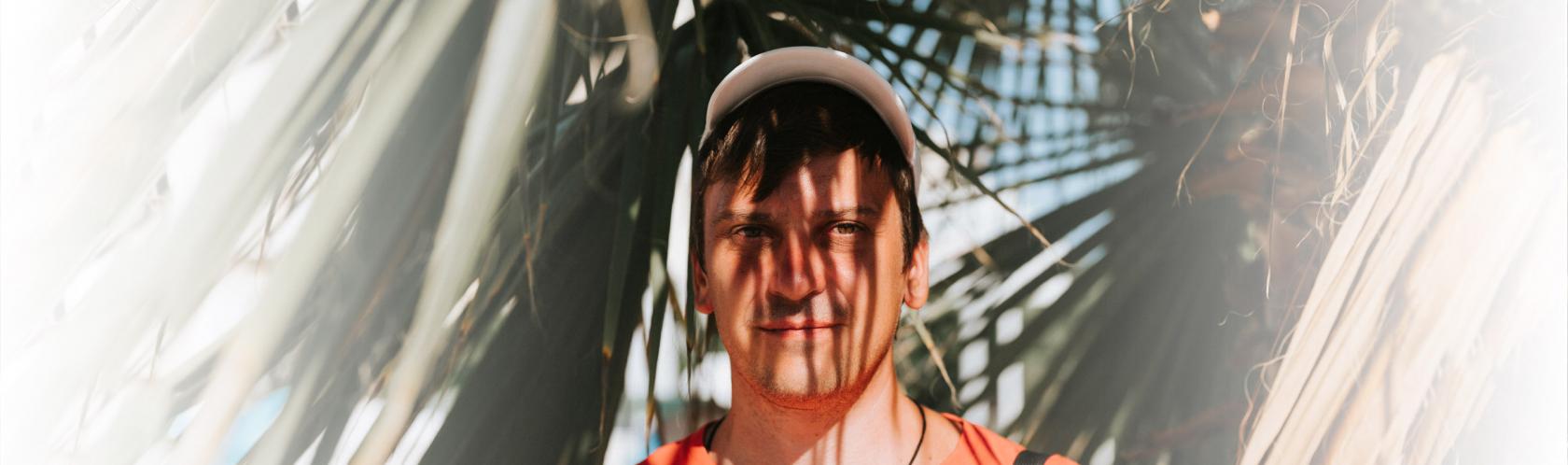 Mężczyzna w czpace z daszkiem w cieniu rzucanym przez liście palmy. Widok twarzy i liści.