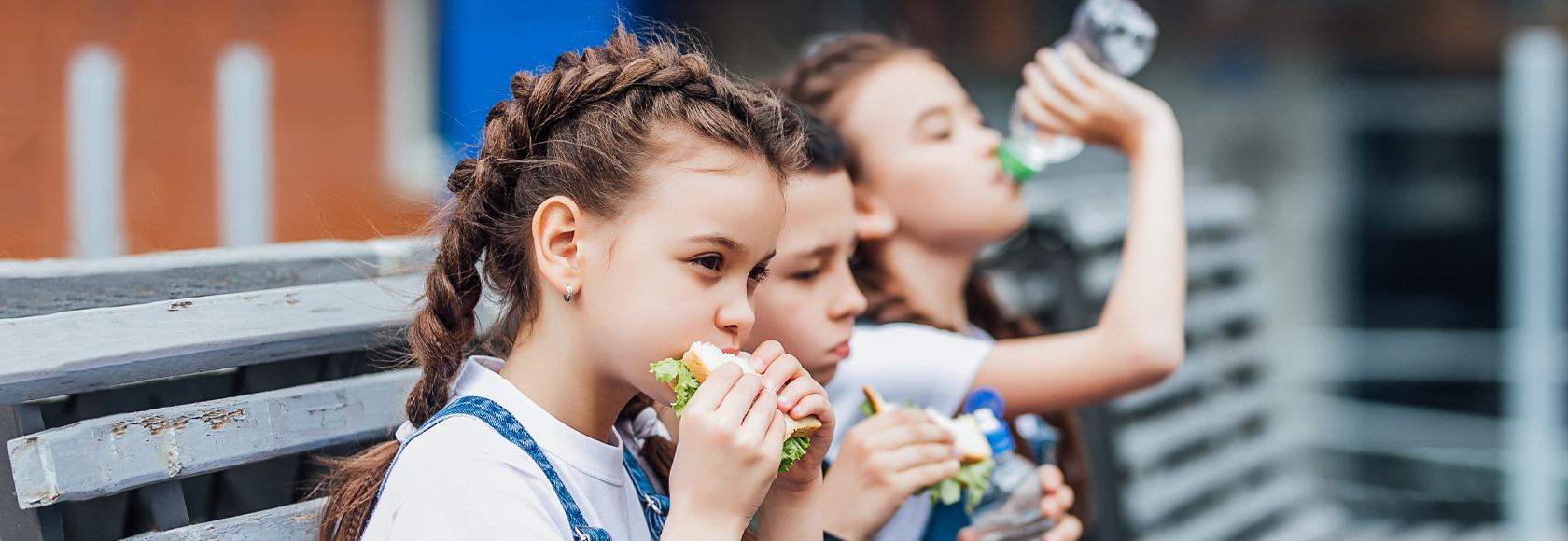 Trójka dzieci siedzi na ławce przed szkołą i je drugie śniadanie. Na pierwszym planie dziewczyna z kanapką z której wystaje sałata, w tle inna pije wodę.