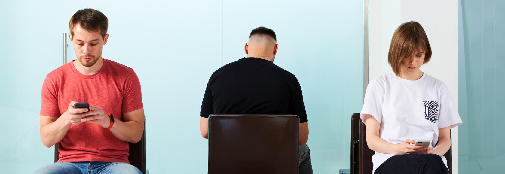 Trzy osoby siedzą na krzesłach w poczekalni w przychodni. Patrzą w telefony.