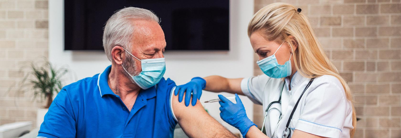 Straszy mężczyna w maseczce siedzący na fotelu. Młoda bobieta w białym fartuchu podaje mu szczepionkę w ramię.