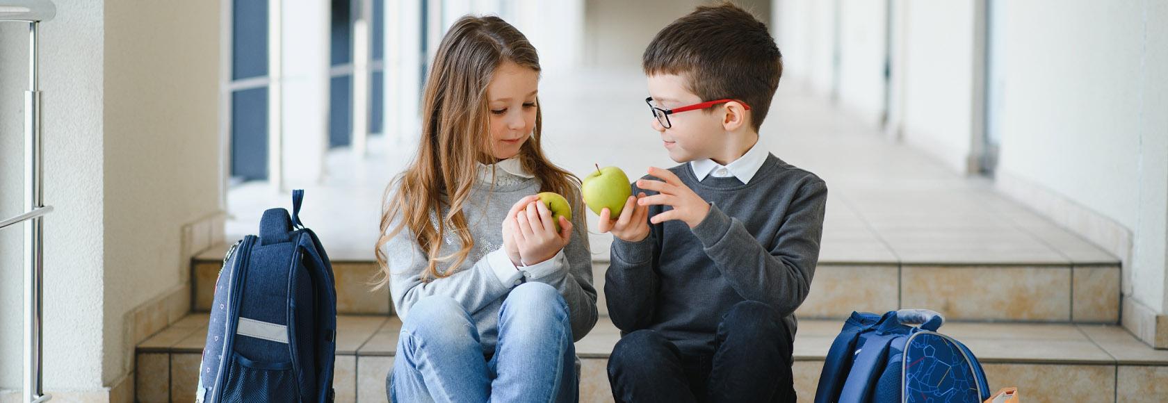 Dziewczynka i chłopiec z podstawówki siedzą na schodach w szkole, trzymają zielone jabłka i patrzą na nie.