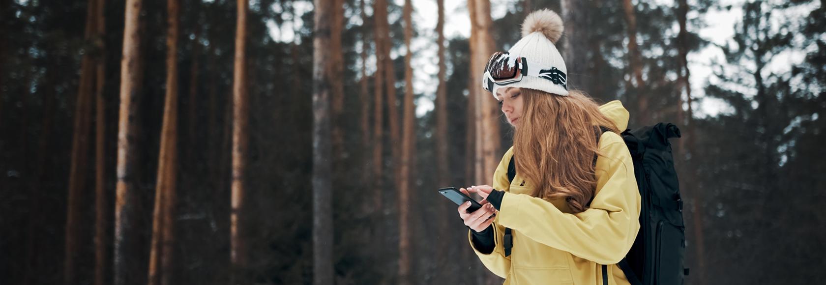 Dziewczyna w żółtej kurtce i czapce na tle lasu. Sprawdza coś w telefonie.