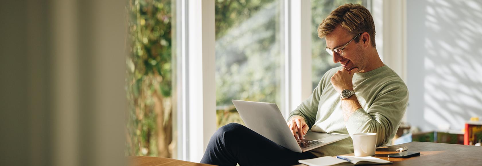 mężczyzna siedzi przy oknie balkonowym z laptopem na kolanach