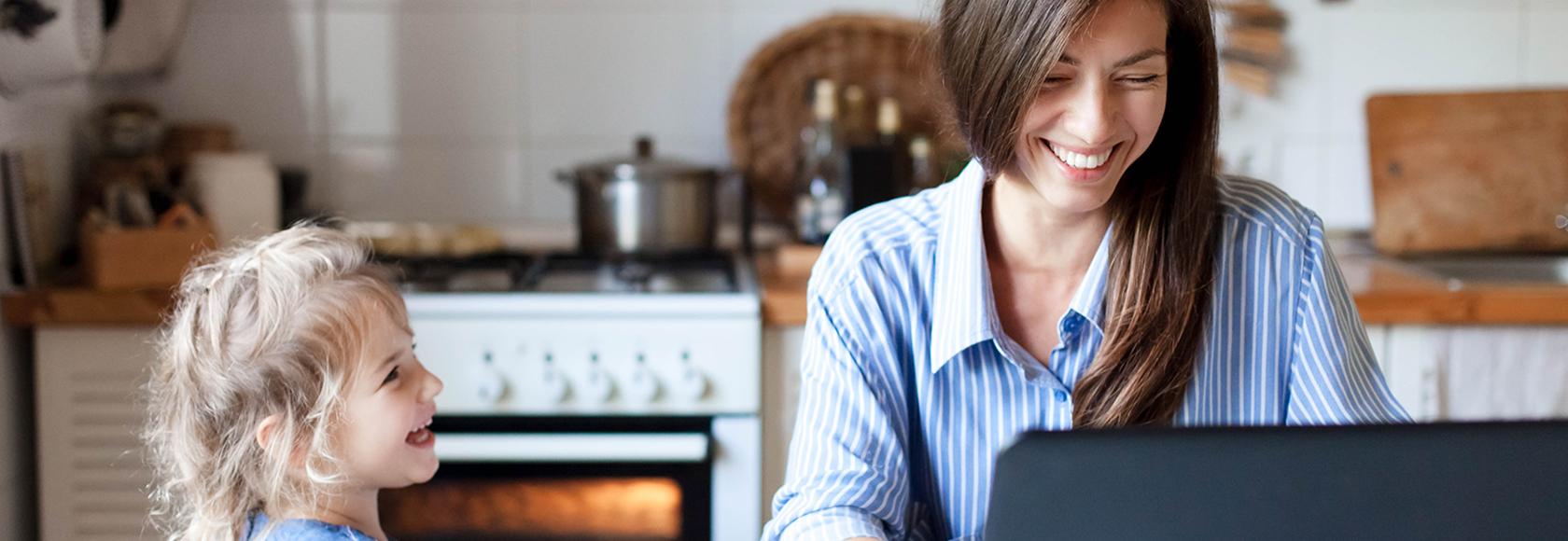 Uśmiechnięta matka przed laptopem, obok smieje się kilkuletnia córka