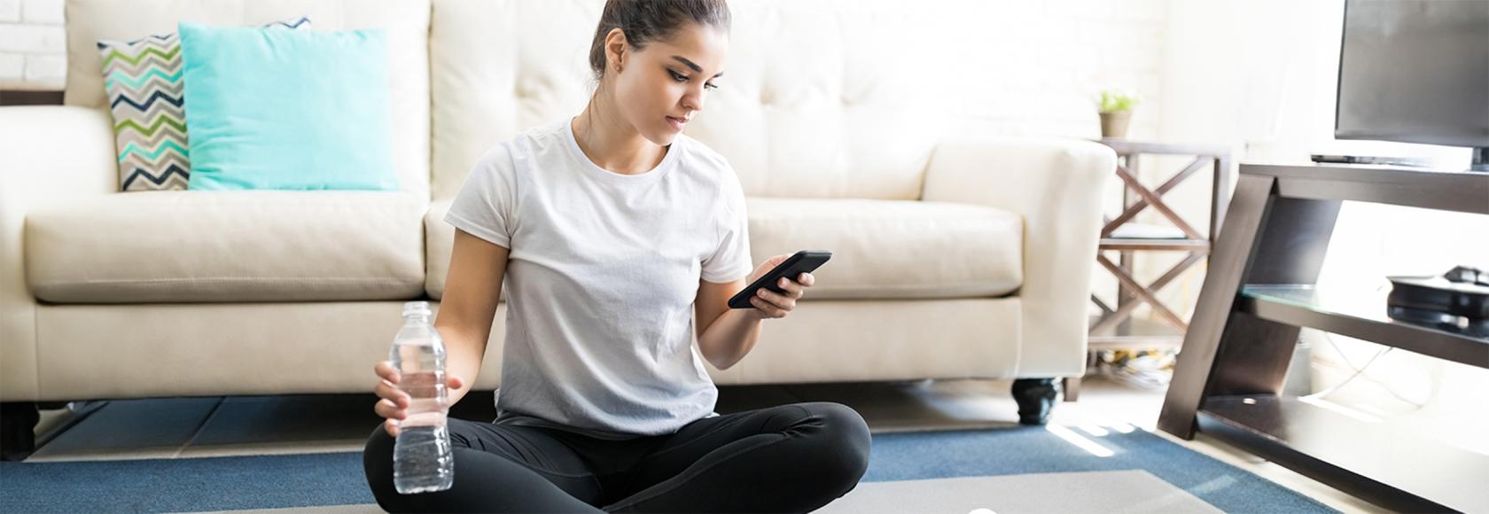 dziewczyna w bałym t-skircie siedzi po turecku w domu, ma w ręku butelkę wody, w drugiej komórkę