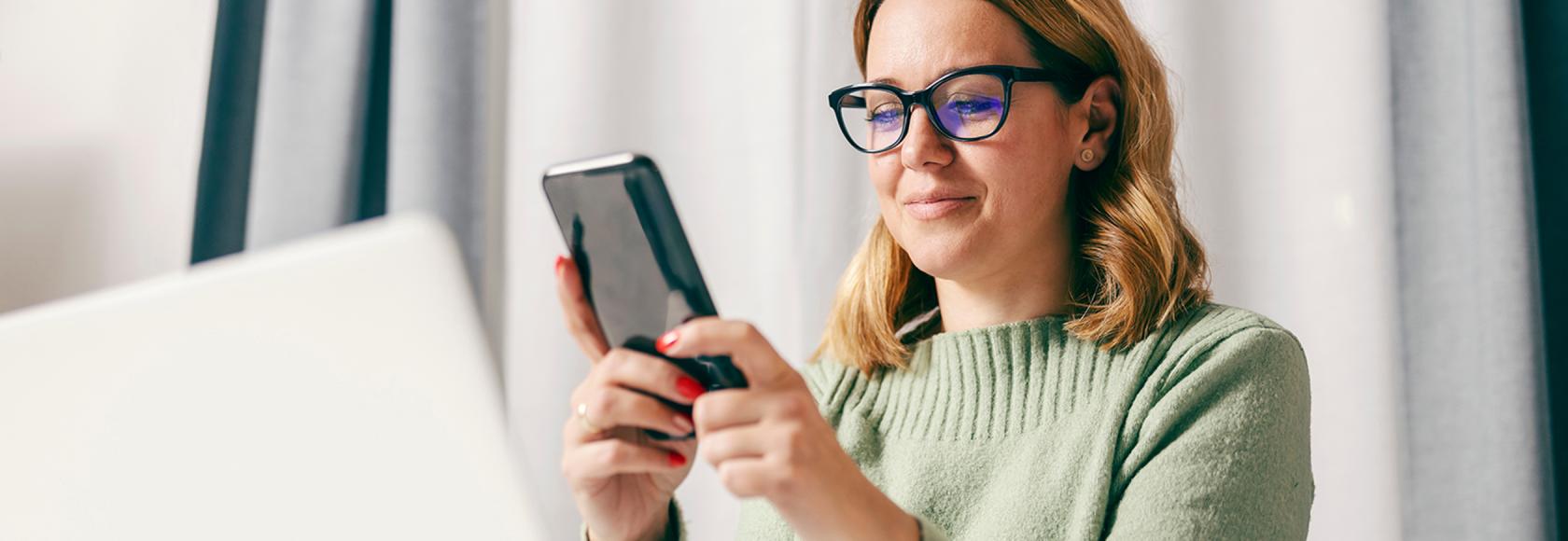 30latka z komórką, na którą patrzy, przed nią laptop