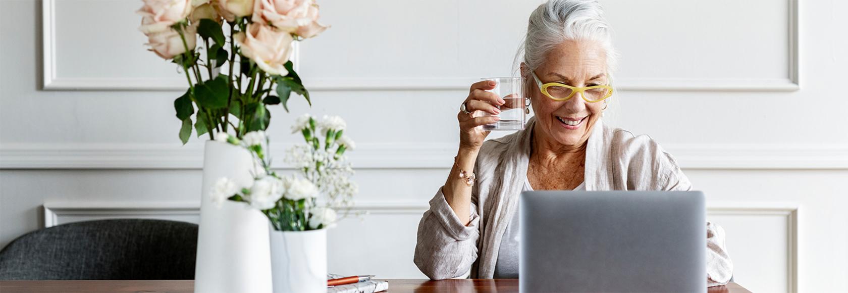 Starsza pani w koku przed laptopem, na stoliki w wazonach dwa bukiety: róże i goździki