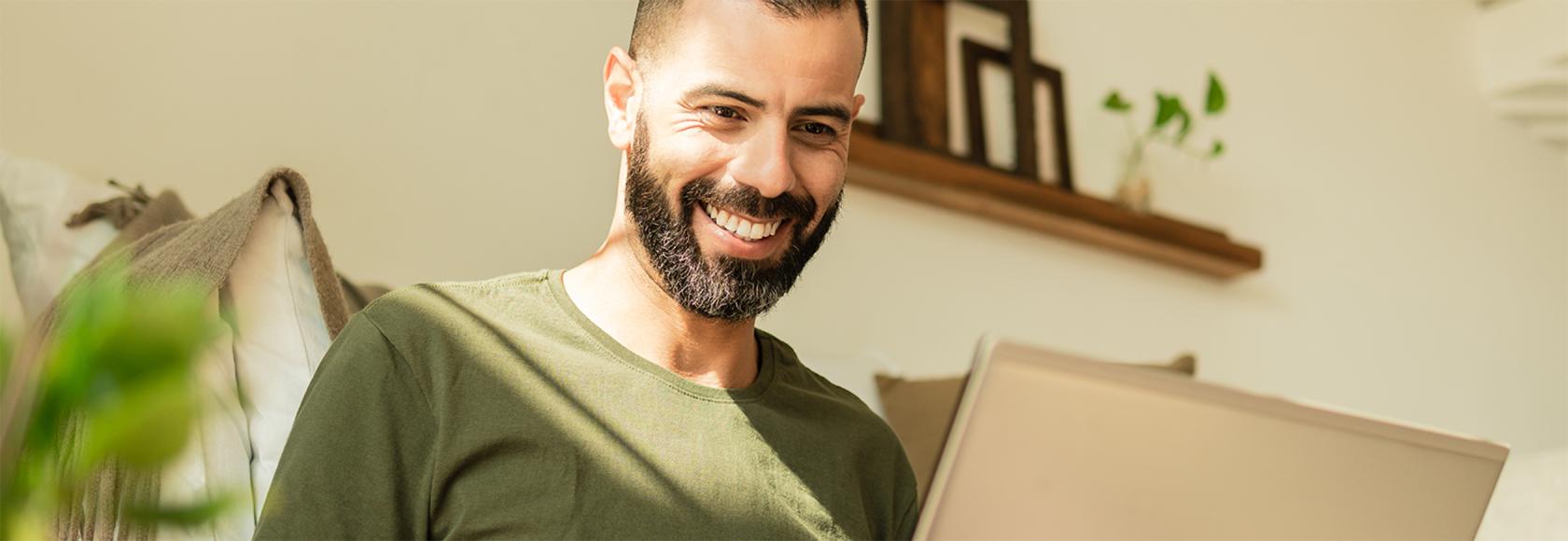 brodaty mężczyzna z laptopem siedzi, za nim widać półkę z wazonem