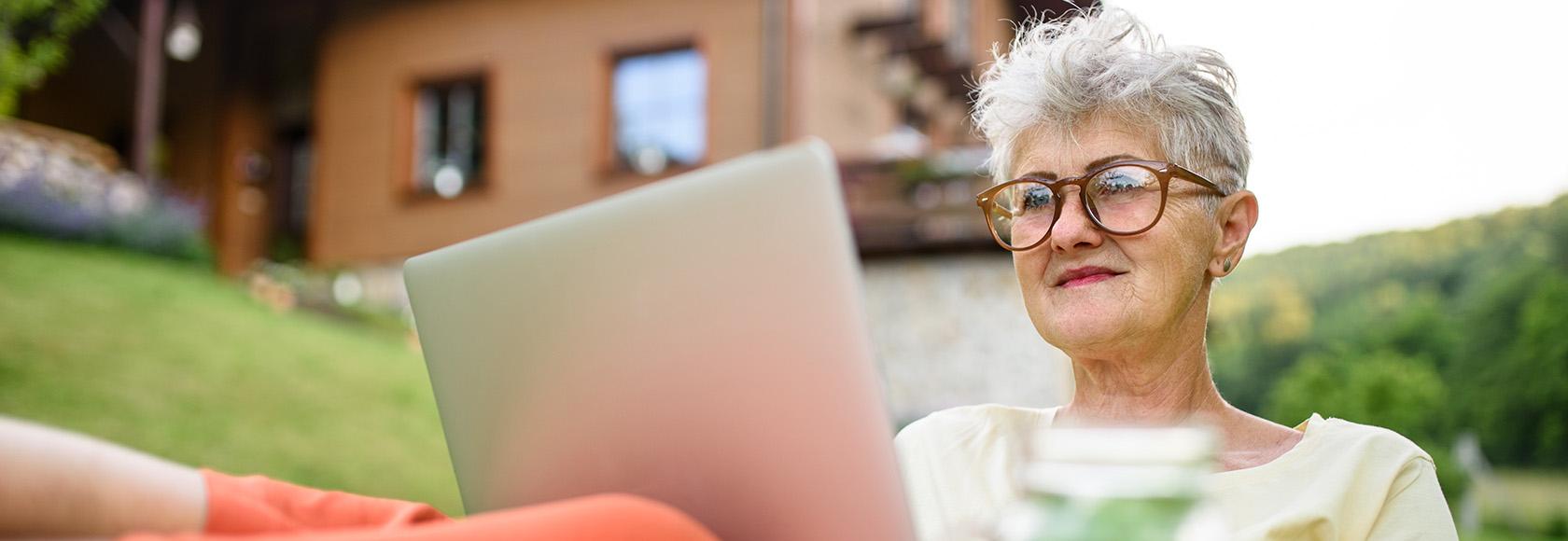 Siwa pani z laptopem przed domem w górach, laptop na kolanach, nogi na stole, na stole arbuz, w tle dom na stoku