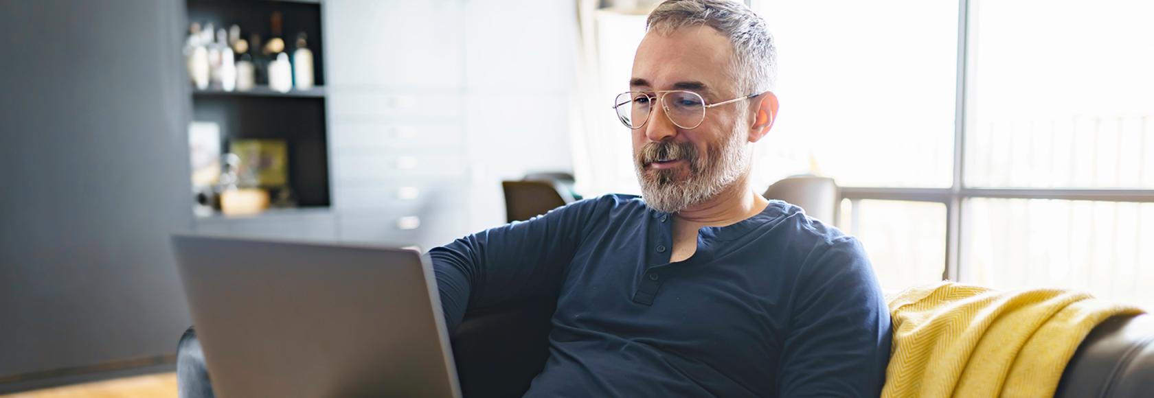 Pan z siwą brodą na kanapie z laptopem na kolanach