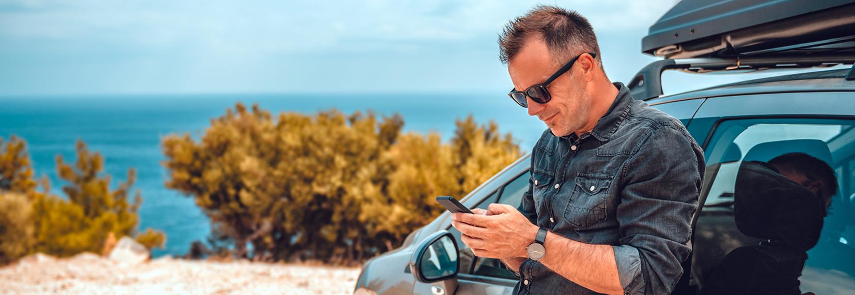 Mężczyzna oparty o auto, z telefonem, w tle krajobraz środziemnomorski
