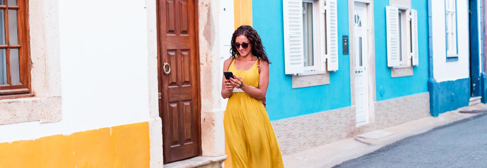 kobieta w żółtej sukience na tle biało-żółtego i bialo-niebieskiego domu, z komórką