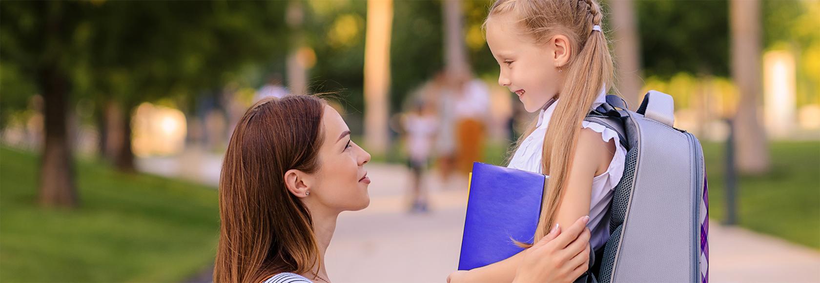 mama kuca i trzyma za ręce córkę z tornistrem, patrza na siebie z uśmiechami
