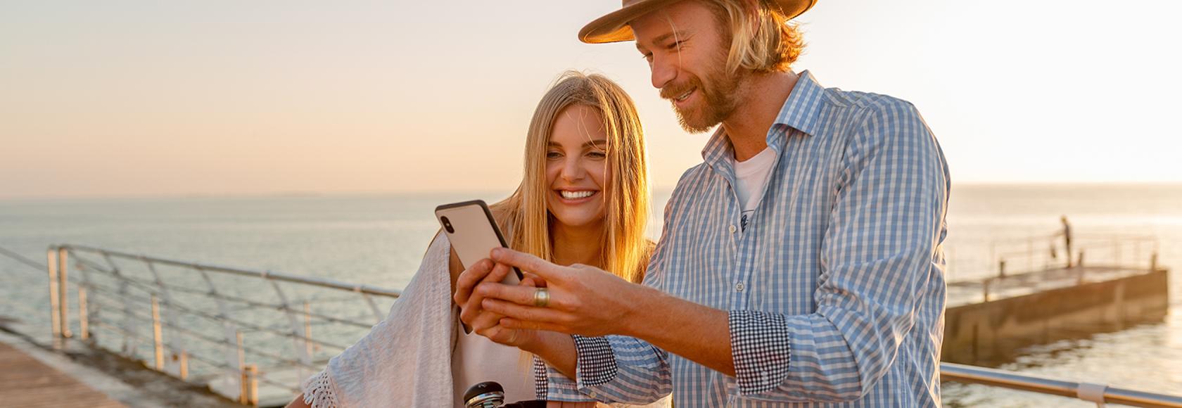 Dziewczyna i chłopak stoją na molo . Dziewczyna trzyma oburącz kierownicę roweru. Mężczyzna pokazuje jej coś na telefonie. W tle morze.