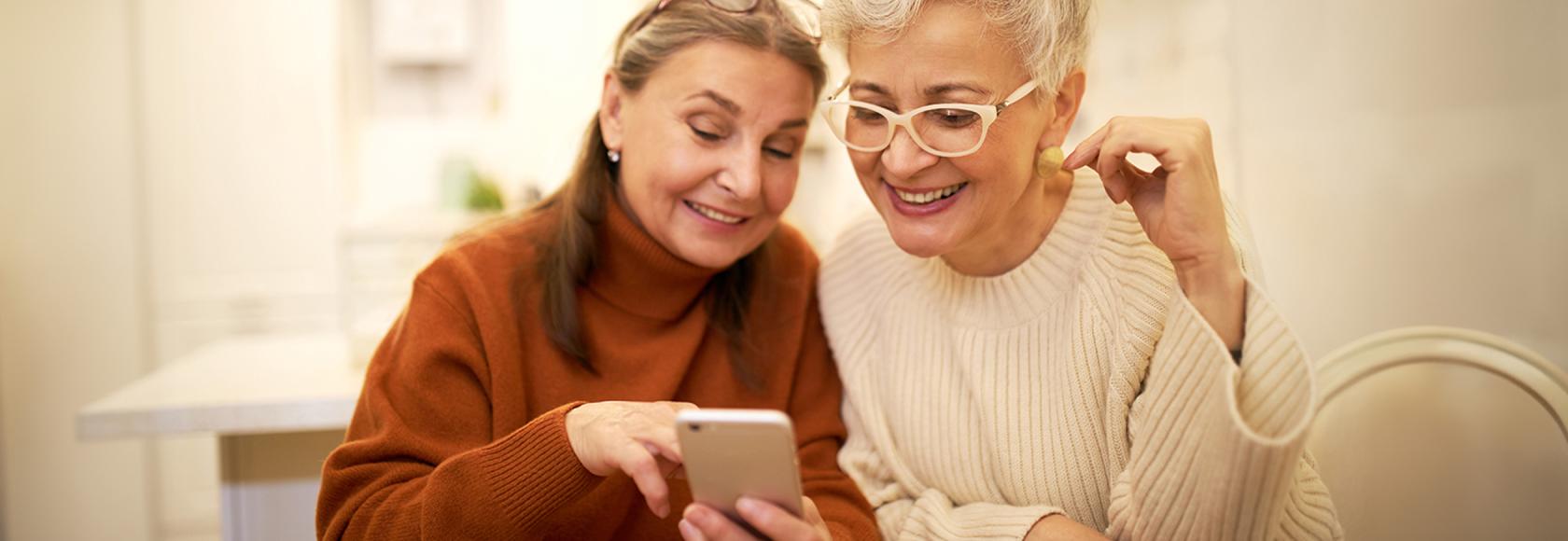 dwie panie w średm wieku w swetrach, jedna pkazuje drugiej coś na komóce