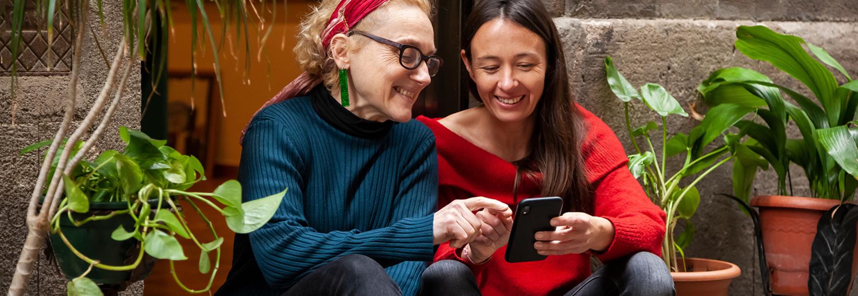 Dwie kobiety siedzą na schodach wśród zieleni i sprawdzają coś na telefonie.