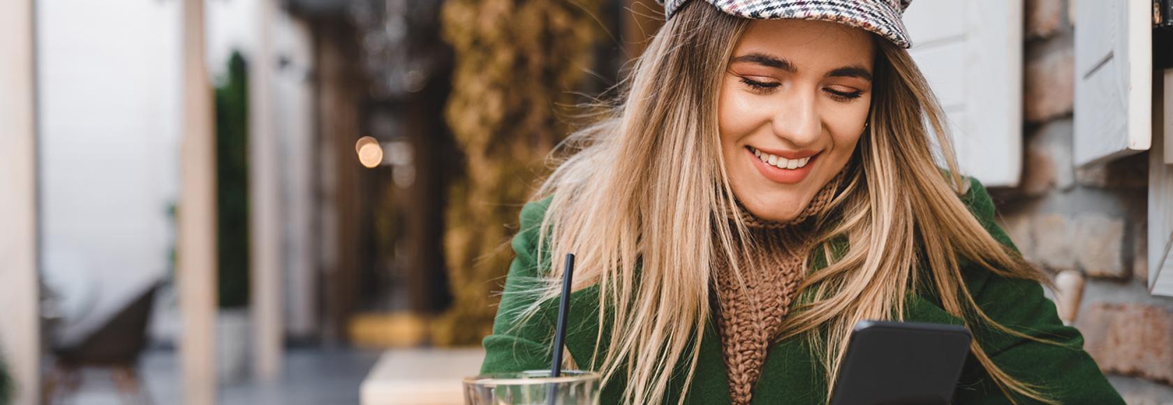 diewczyna w kyrtce i czapce z daszkiem pije kawę przy stoliku na ulicy i patrzy w komórkę