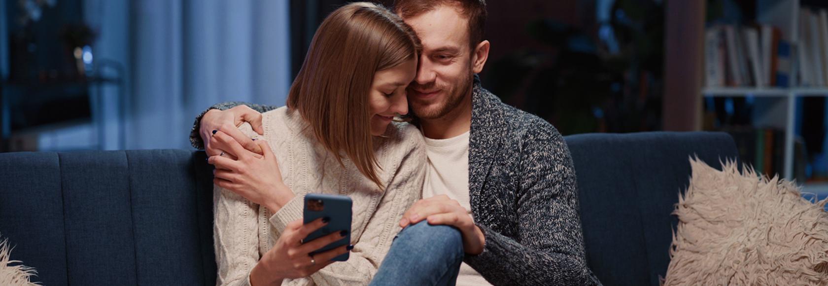 Przytulona para na kanapie, ona trzyma w ręku komórkę