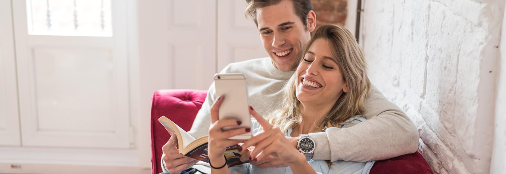 para siedzi wtulona na kanapie, on z ksiazka, ona z komórką mu coś pokazuje z uśmiechem