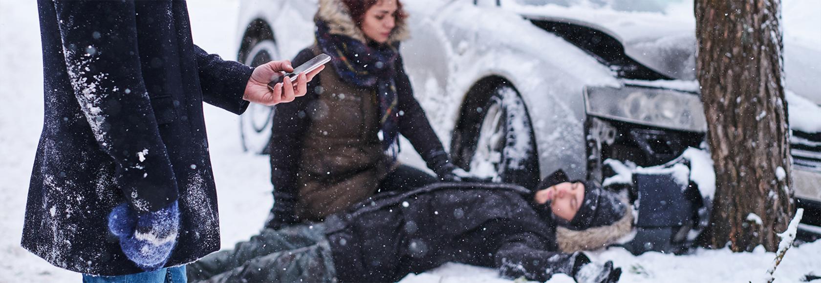 śnieg, wypadek samochodowy, jedna osoba trzyma komorke, druga udziela pomocy poszkodowanemu
