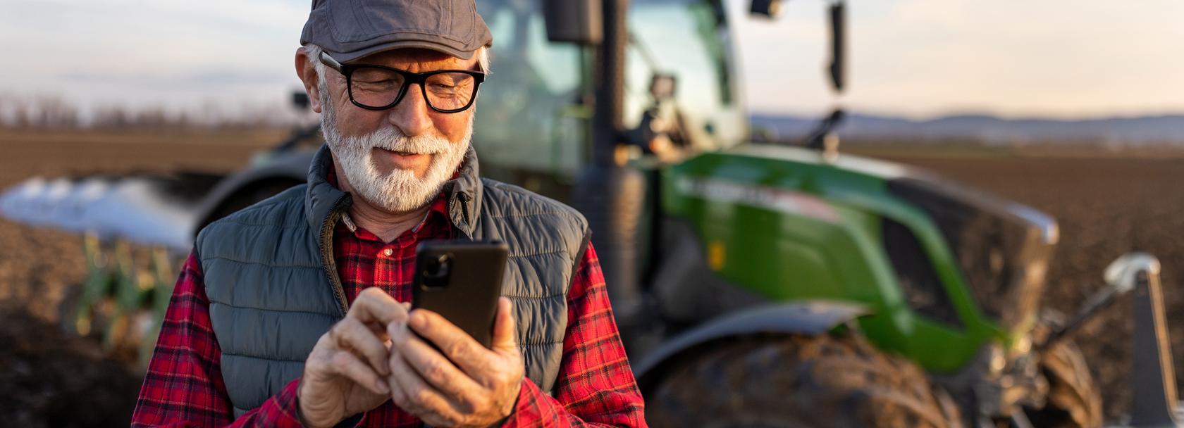 Pan z siwą brodą z komórką, w tle traktor