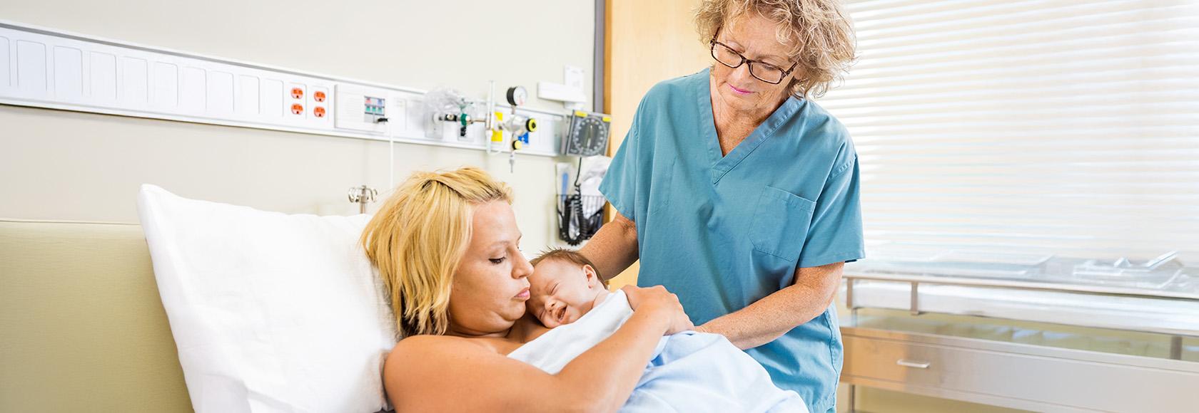 Kobieta leży z noworodkiem, stoi położna