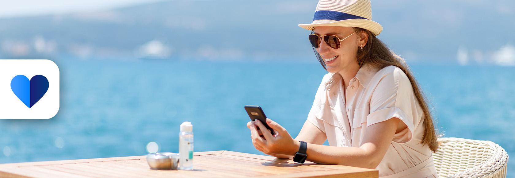 Kobieta siedzi prrzy stoliku, na stole butelka wody, ona w kapeluszu i ciemnych okularach, z komórką, w tle morze