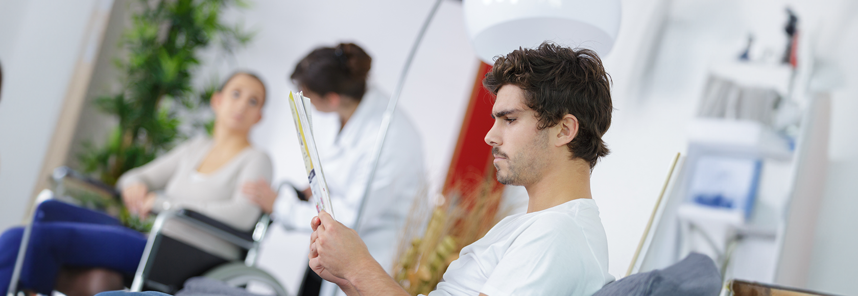 Młody mężczyzna czyta broszurę informacyjną. W tle osoba na wózku inwalidzkim i lekarka.