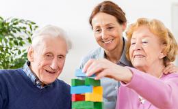 Starsi państwo ćwiczą układankę pod okiem opiekunki