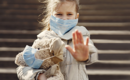 Dziewczynka z misiem pod ręką, mają założone maseczki. Lewa ręka dziewczynki wyciągnięta w przód, otwartą dłonią.