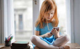 Dziewczynka siedząca ze skrzyżowanymi nogami na tle okna, przeglądająca telefon komórkowy.