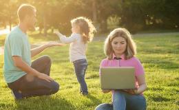 Ojciec bawi się z córeczką na łące, matka zagląda do laptopa